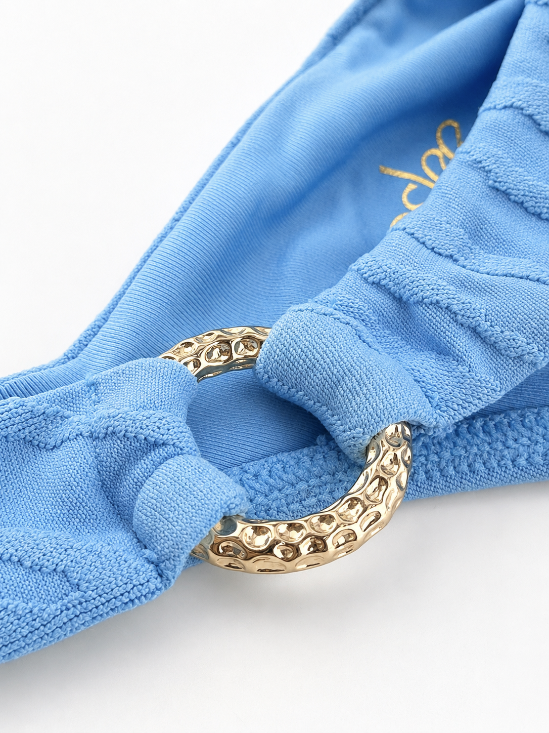 Close-up of a light blue textured fabric swim bikini bottom with a large hammered gold metal ring detail, shown on a white background.