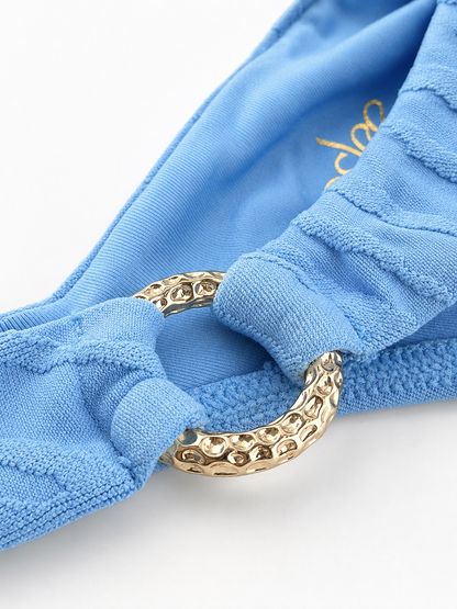 Close-up of a light blue textured fabric swim bikini bottom with a large hammered gold metal ring detail, shown on a white background.
