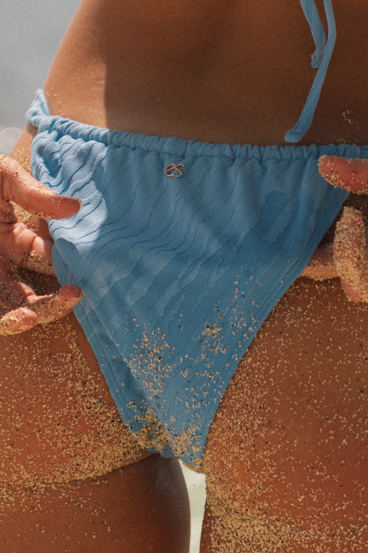 A close-up of a person wearing a light blue textured bikini bottom on a sandy beach, with sand scattered across the fabric and skin.