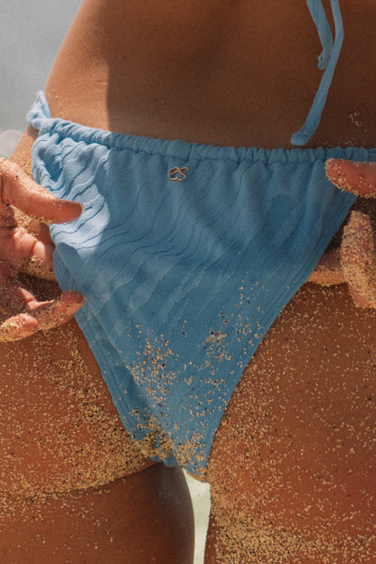 A close-up of a person wearing a light blue textured bikini bottom on a sandy beach, with sand scattered across the fabric and skin.