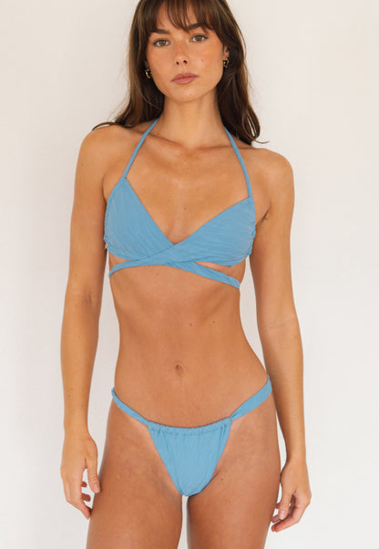 A woman modeling a light blue two-piece bikini with a halter top and matching low-rise bottoms against a plain off-white background.