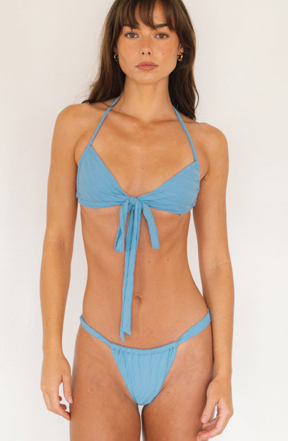 A woman modeling a light blue two-piece bikini with a halter top and matching side-tie bottoms against a plain white studio background.