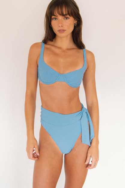 A woman modeling a light blue two-piece bikini with an underwire bikini top and side-tie high-waisted bottoms, set against a plain off-white background.