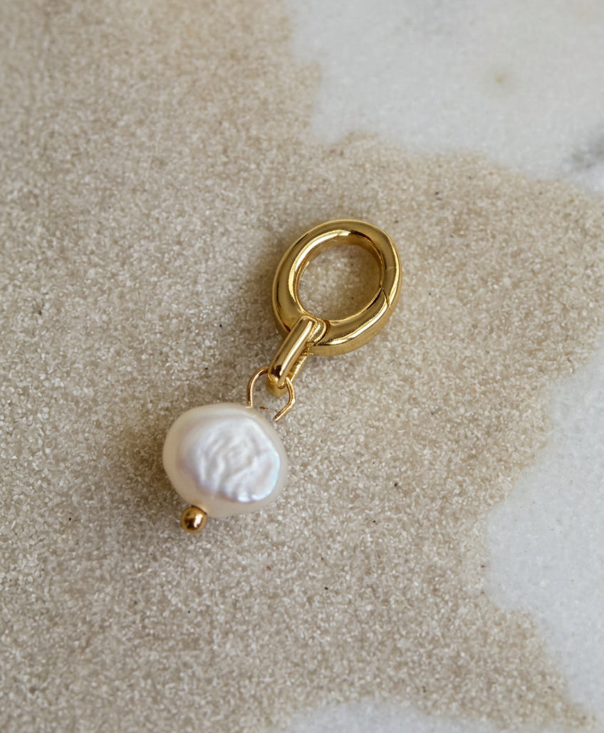 A gold-tone bikini charm featuring a single baroque pearl pendant, displayed on a textured beige stone surface.