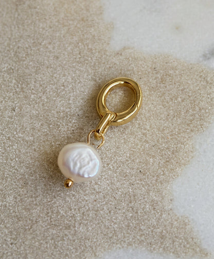A gold-tone bikini charm featuring a single baroque pearl pendant, displayed on a textured beige stone surface.