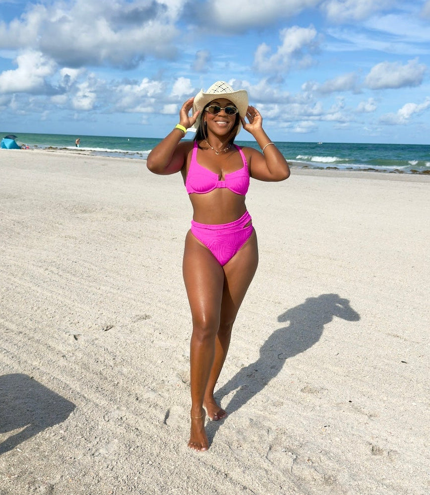 A woman wearing a bright pink two-piece bikini walks on a sandy beach under a blue sky with white clouds, holding a beige straw hat.