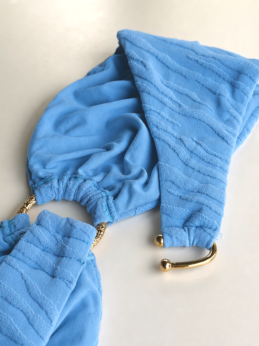 A close up detail of a light blue textured swimsuit bikini top with a gold metal ring and gold metal U-hook back closure.