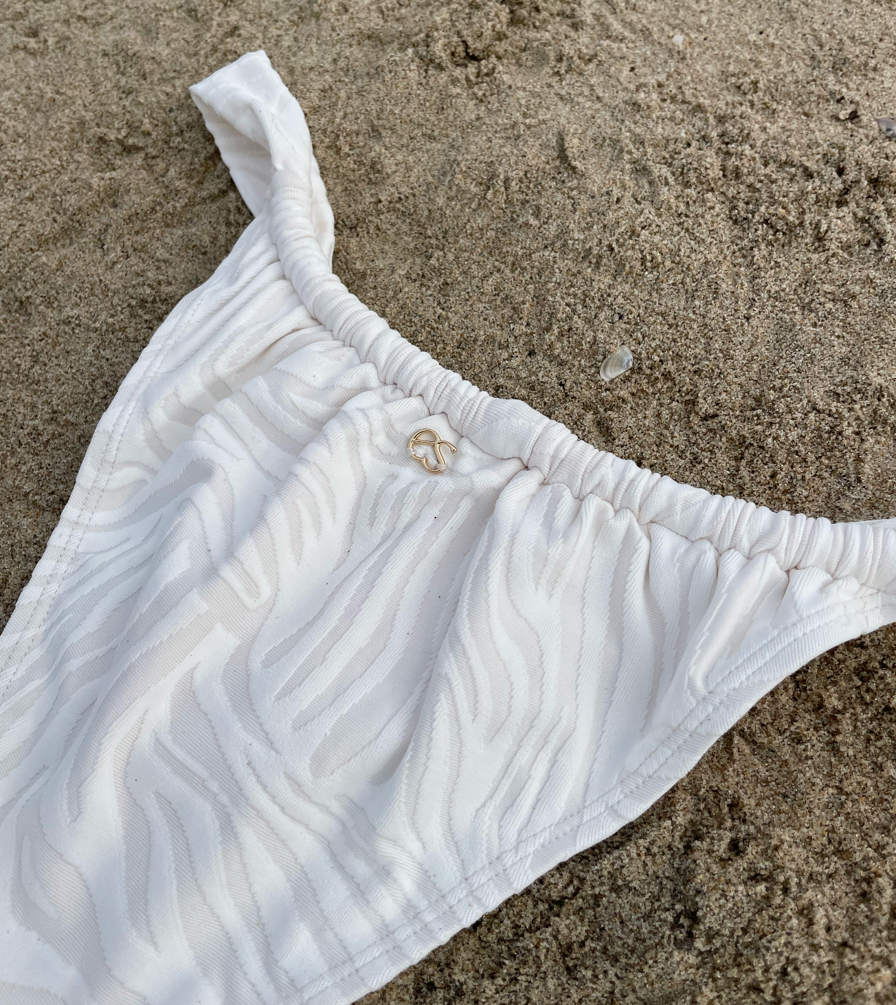 White bikini bottom with zebra texture lying on the sand with gold logo charm